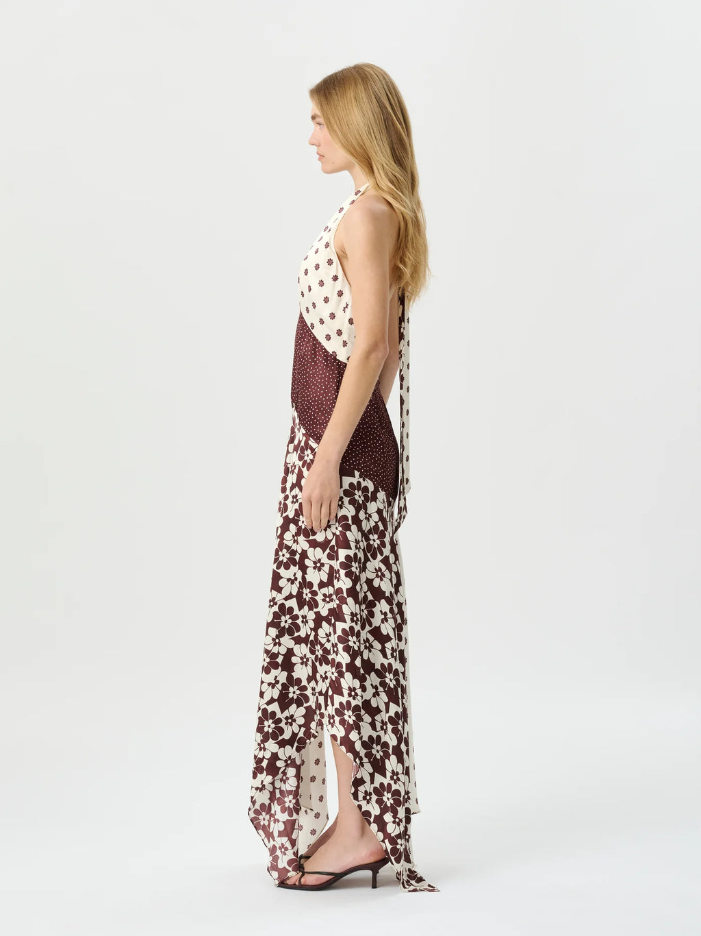 A woman stands in profile wearing a sleeveless maxi dress with a cream base, dark red floral print on the skirt, and a dark red mesh bodice with a white polka-dot upper panel, photographed against a plain light gray studio background.