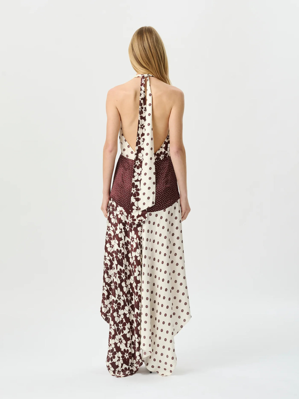A woman stands with her back to the camera wearing a halter-neck maxi dress with a split design: one side has a dark red and cream floral print, and the other side has a dark red and cream polka-dot print.