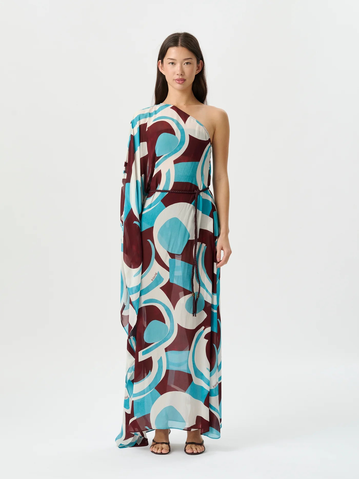 A model stands against a plain white studio background wearing a one-shoulder maxi dress with a bold abstract geometric pattern in teal, burgundy, cream, and white.