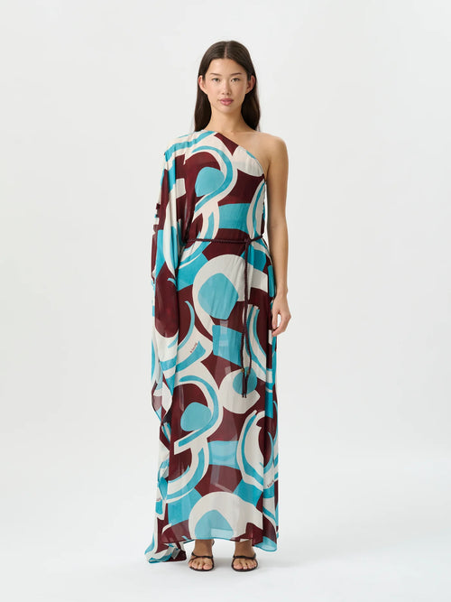 A model stands against a plain white studio background wearing a one-shoulder maxi dress with a bold abstract geometric pattern in teal, burgundy, cream, and white.