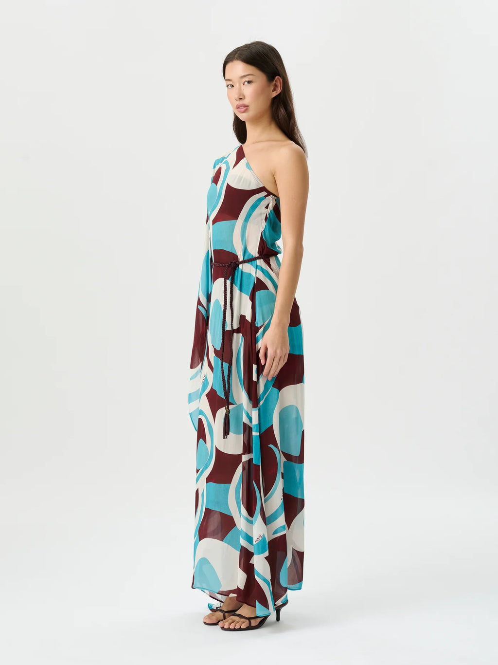 A woman modeling a sleeveless maxi dress with an abstract geometric pattern in teal, burgundy, cream, and brown against a plain light gray studio background.