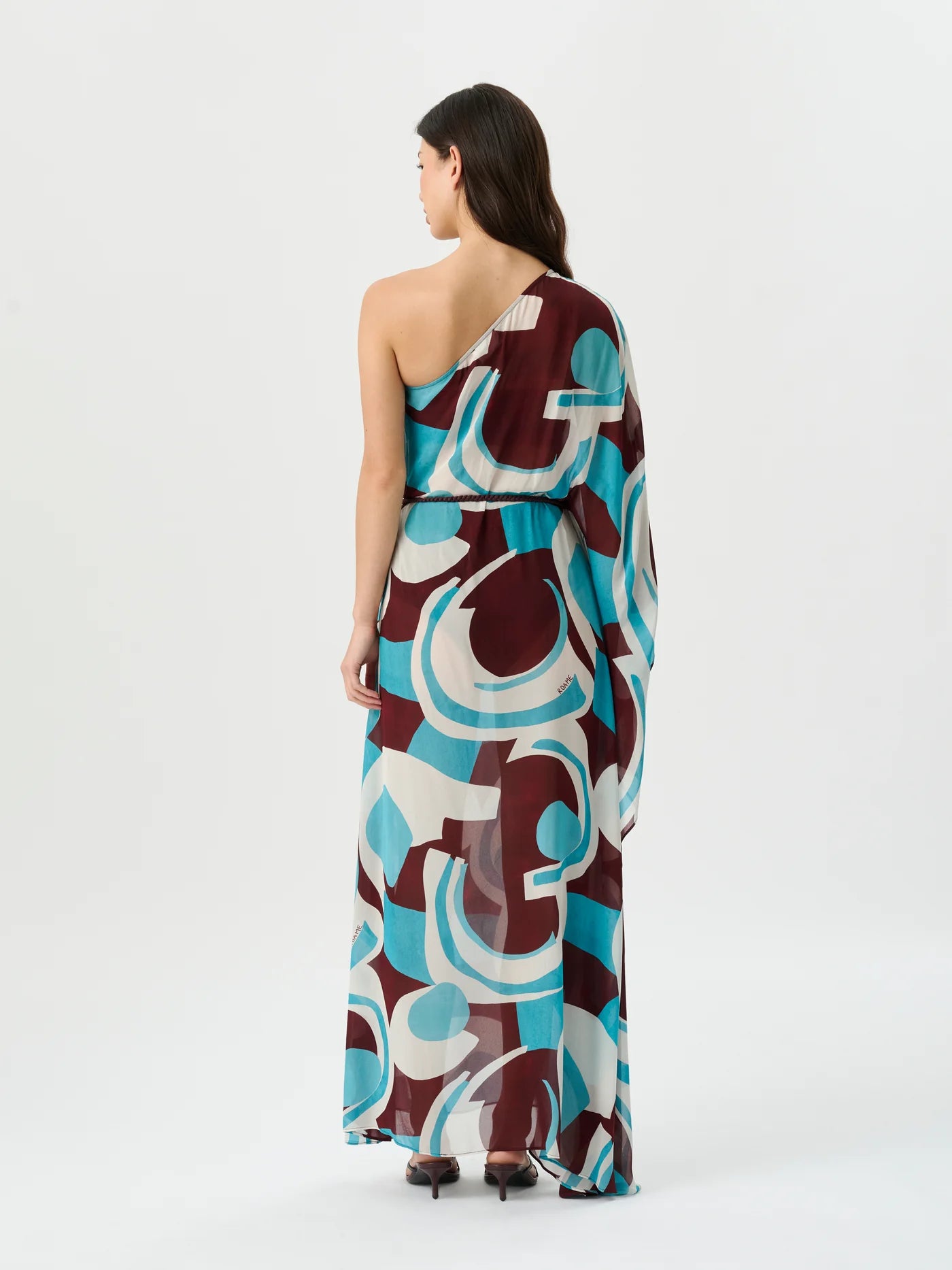 A woman viewed from the back wearing a one-shoulder maxi dress with a bold abstract geometric pattern in teal, burgundy, and off-white, set against a plain white studio background.