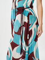 A close-up of a sleeveless dress with an abstract geometric pattern in teal, burgundy, and off-white, featuring a dark brown braided tie belt at the waist.