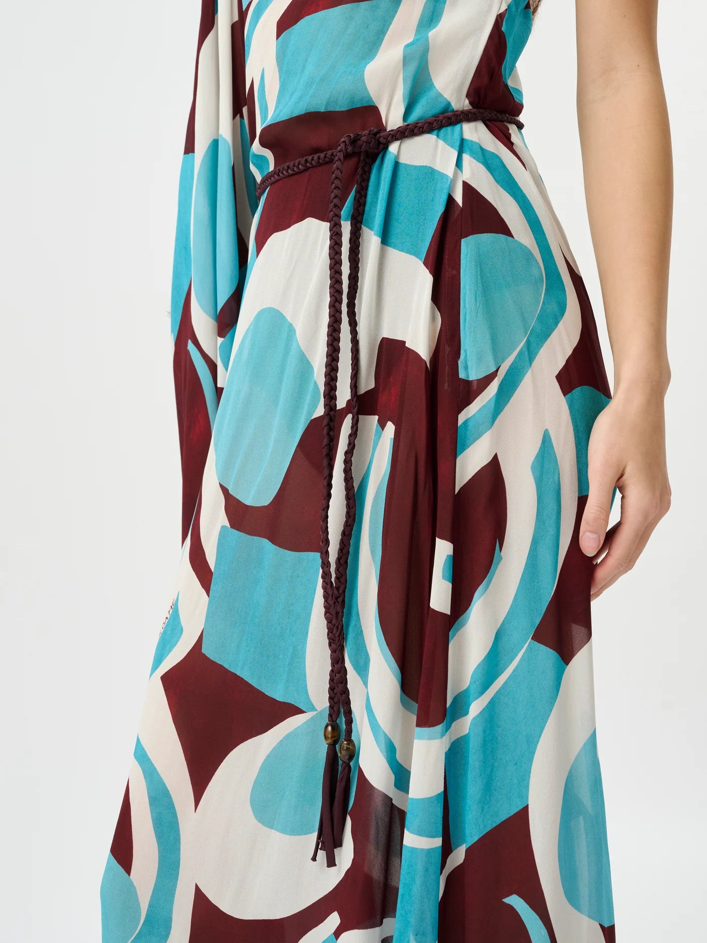 A close-up of a sleeveless dress with an abstract geometric pattern in teal, burgundy, and off-white, featuring a dark brown braided tie belt at the waist.