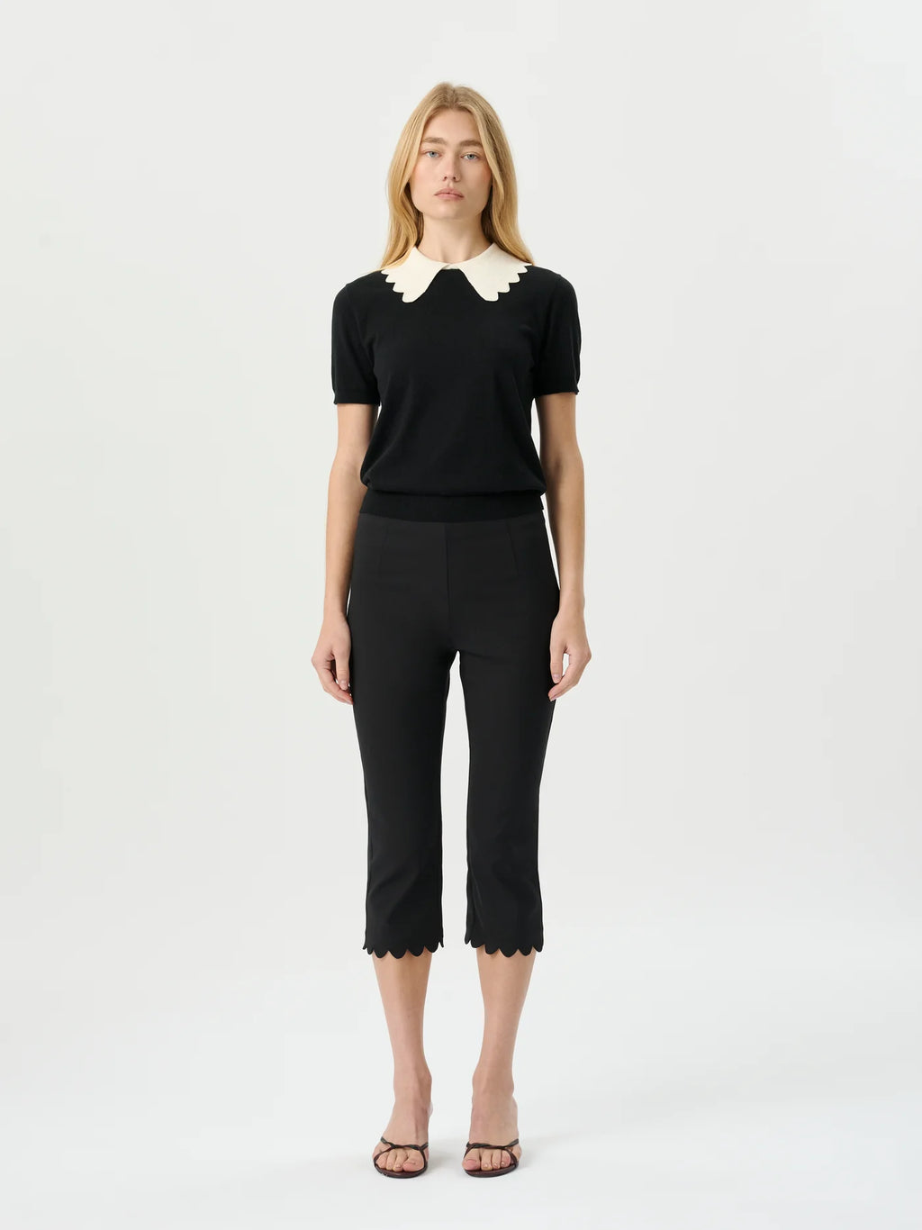 A woman modeling a black short-sleeve knit top with a cream scalloped Peter Pan collar, paired with black cropped trousers and brown strappy sandals, against a plain white studio background.