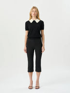 A woman modeling a black short-sleeve knit top with a cream scalloped Peter Pan collar, paired with black cropped trousers and brown strappy sandals, against a plain white studio background.