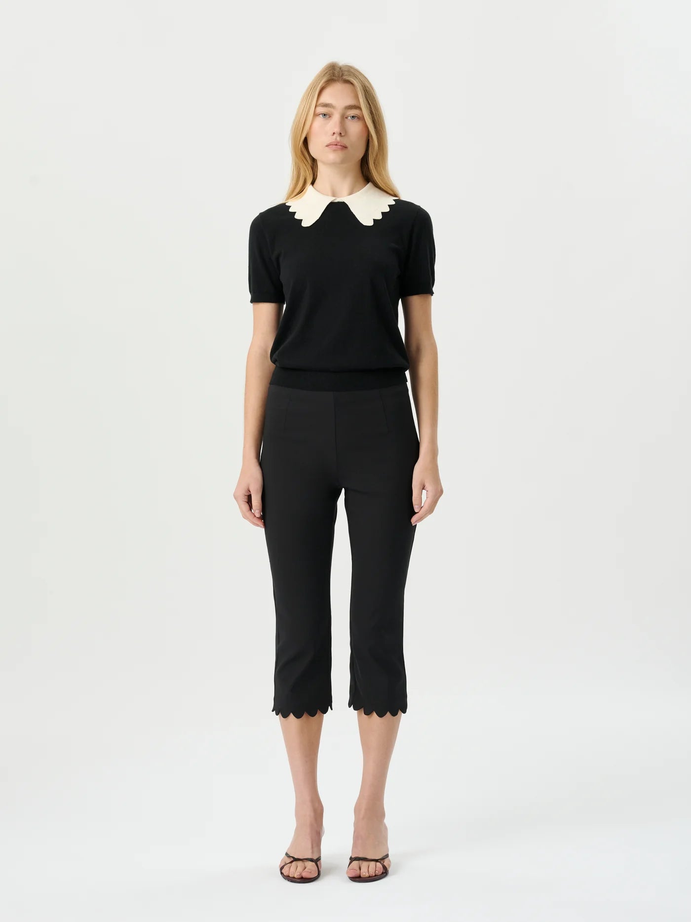 A woman modeling a black short-sleeve knit top with a cream scalloped Peter Pan collar, paired with black cropped trousers and brown strappy sandals, against a plain white studio background.