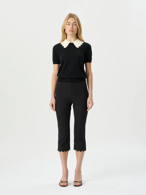 A woman modeling a black short-sleeve knit top with a cream scalloped Peter Pan collar, paired with black cropped trousers and brown strappy sandals, against a plain white studio background.
