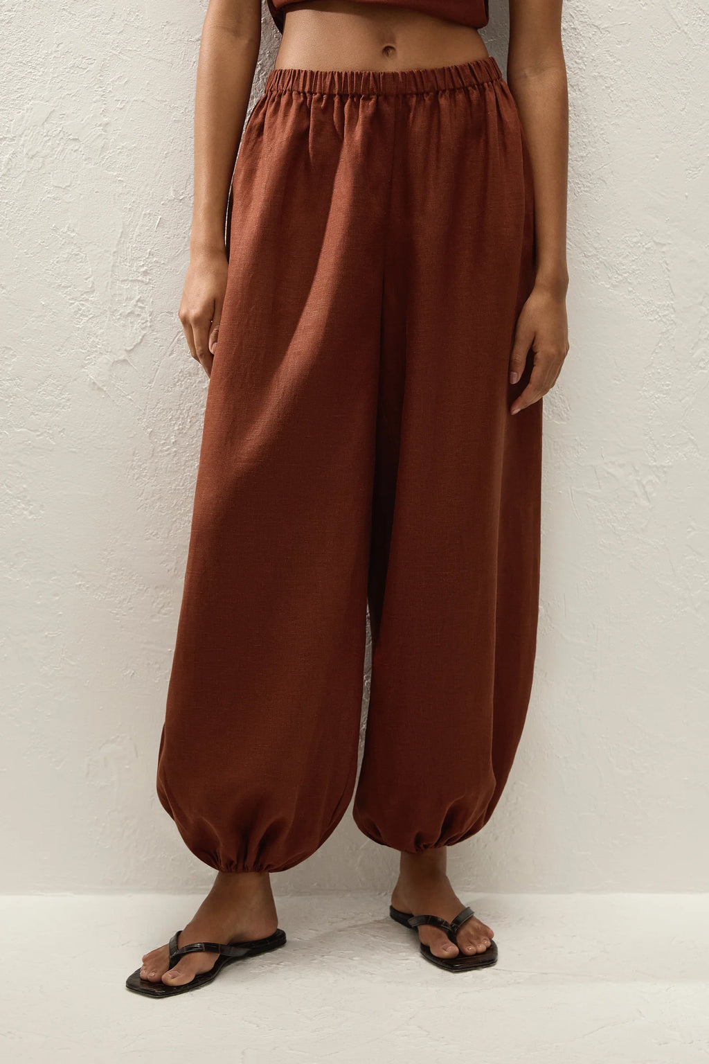 A person wearing loose, wide-leg rust-brown harem-style pants with an elastic waist and gathered cuffs, paired with black flip-flop sandals against a textured off-white wall.