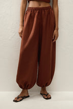 A person wearing loose, wide-leg rust-brown harem-style pants with an elastic waist and gathered cuffs, paired with black flip-flop sandals against a textured off-white wall.