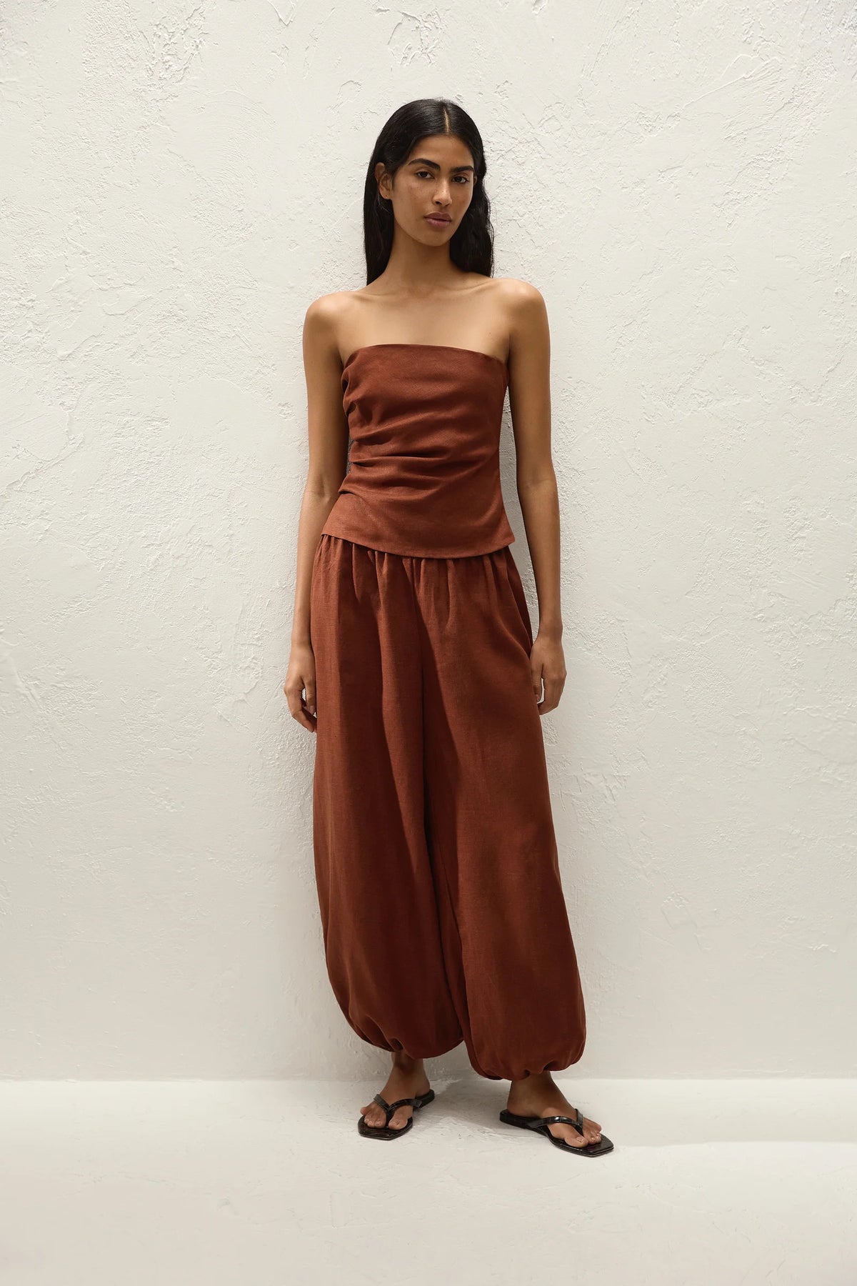 A woman modeling a rust-brown strapless tube top and matching wide-leg harem pants against a textured white wall.