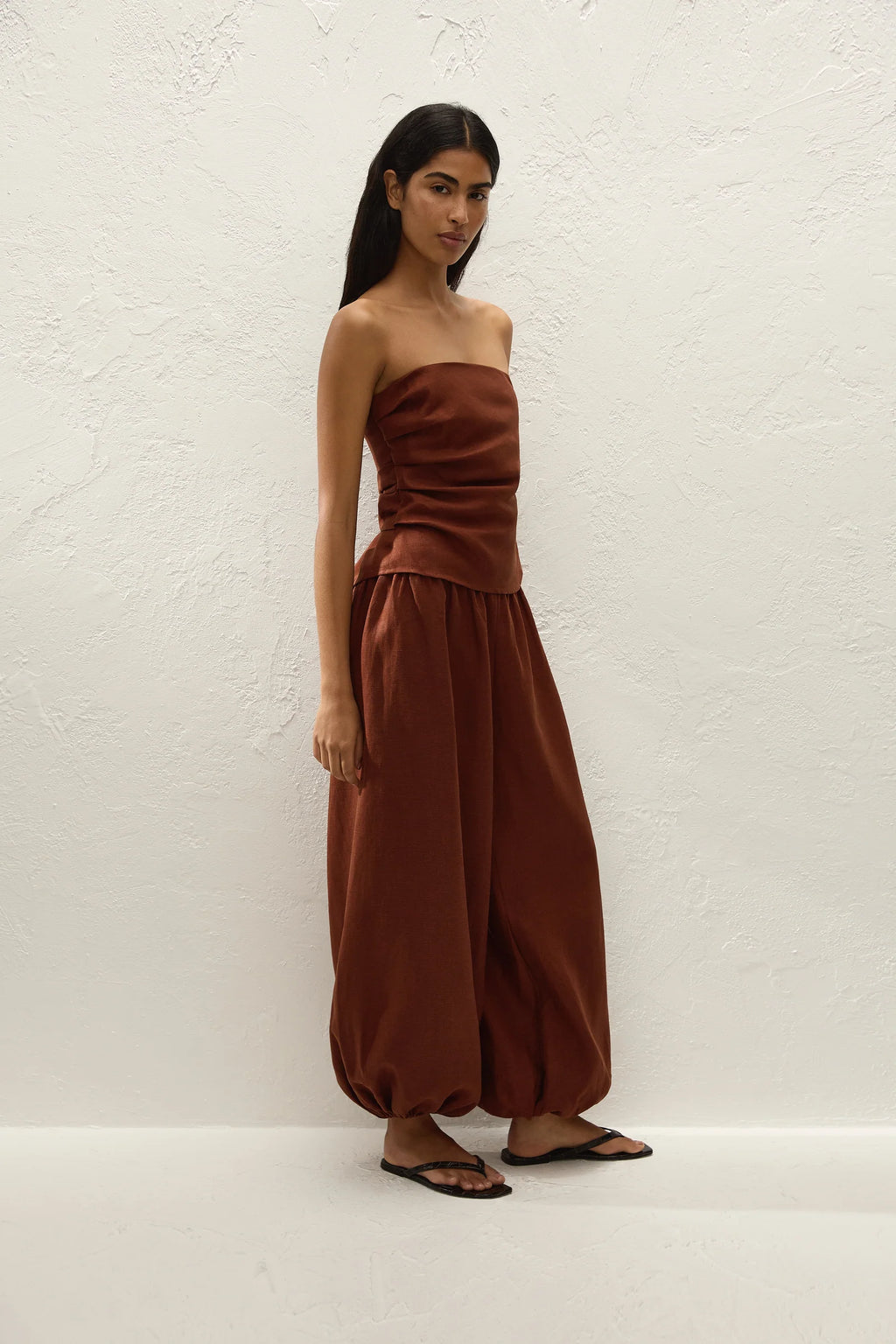 A woman modeling a rust-brown strapless tube top and matching wide-leg harem pants against a textured white wall.