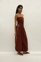 A woman modeling a rust-brown strapless tube top and matching wide-leg harem pants against a textured white wall.