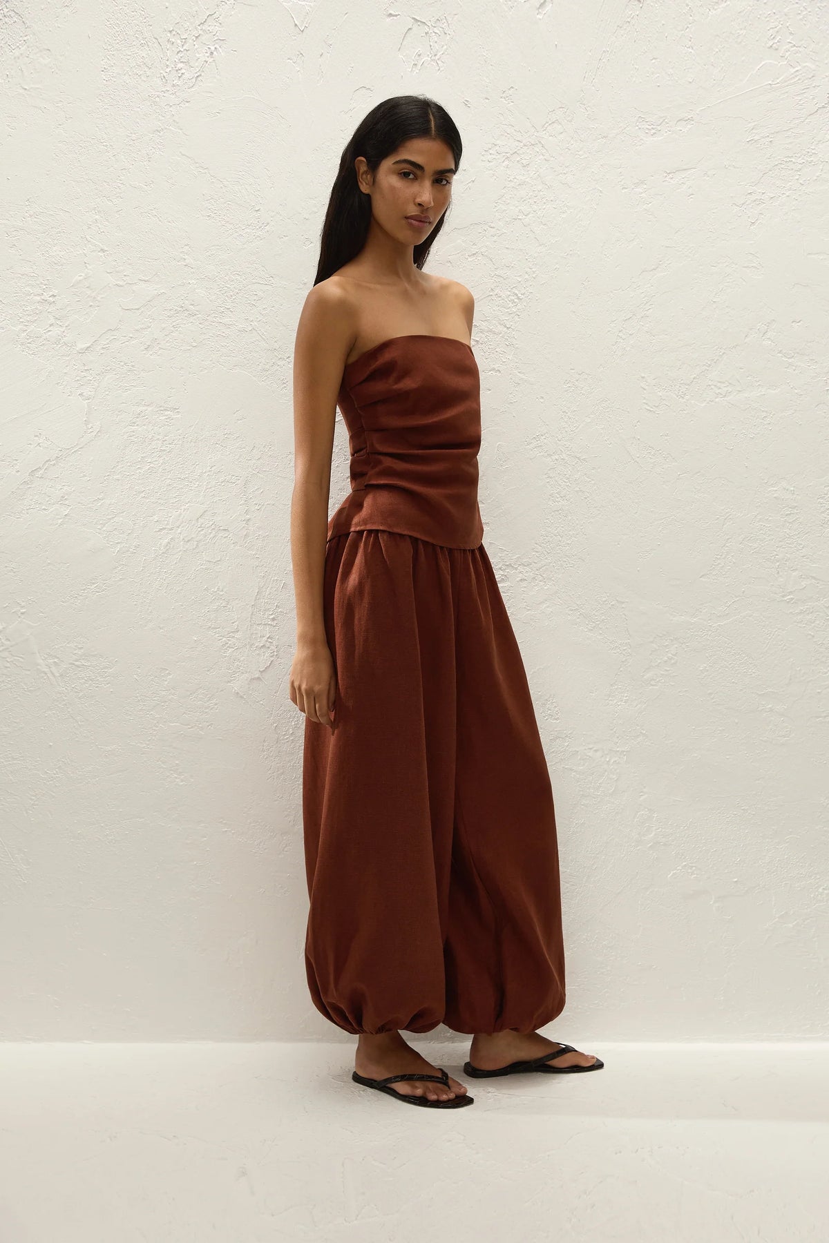 A woman modeling a rust-brown strapless tube top and matching wide-leg harem pants against a textured white wall.