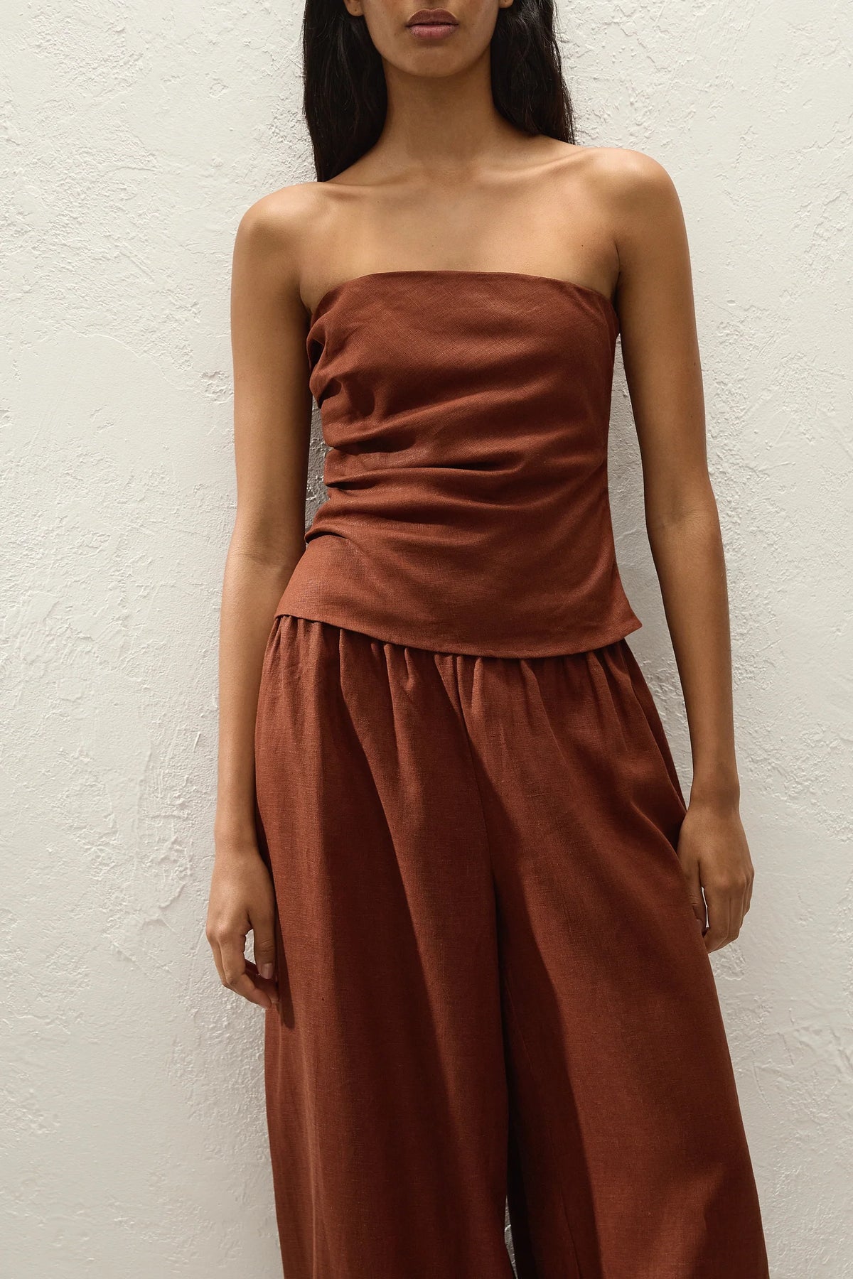 A woman wearing a rust-brown strapless top and matching wide-leg pants, standing against a textured off-white wall.
