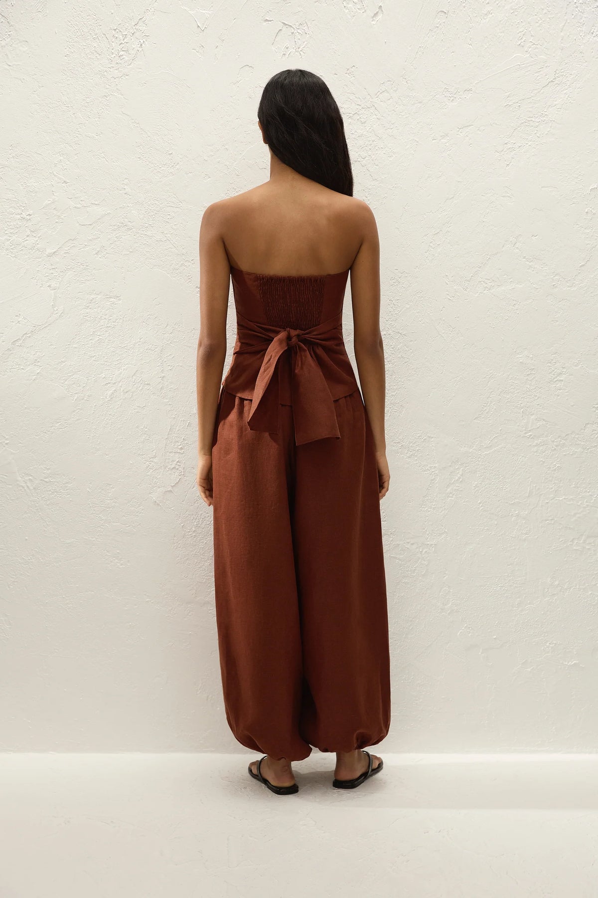 A woman stands with her back to the camera wearing a rust-brown strapless jumpsuit with wide legs and a tied waist detail, set against a textured off-white wall.