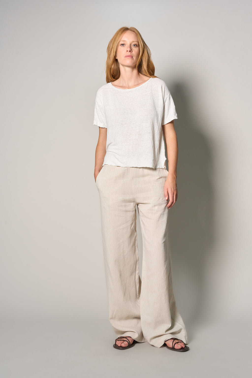 A woman modeling a white short-sleeve crewneck t-shirt and beige wide-leg linen pants, paired with dark sandals, against a neutral light gray studio background.