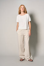 A woman modeling a white short-sleeve crewneck t-shirt and beige wide-leg linen pants, paired with dark sandals, against a neutral light gray studio background.