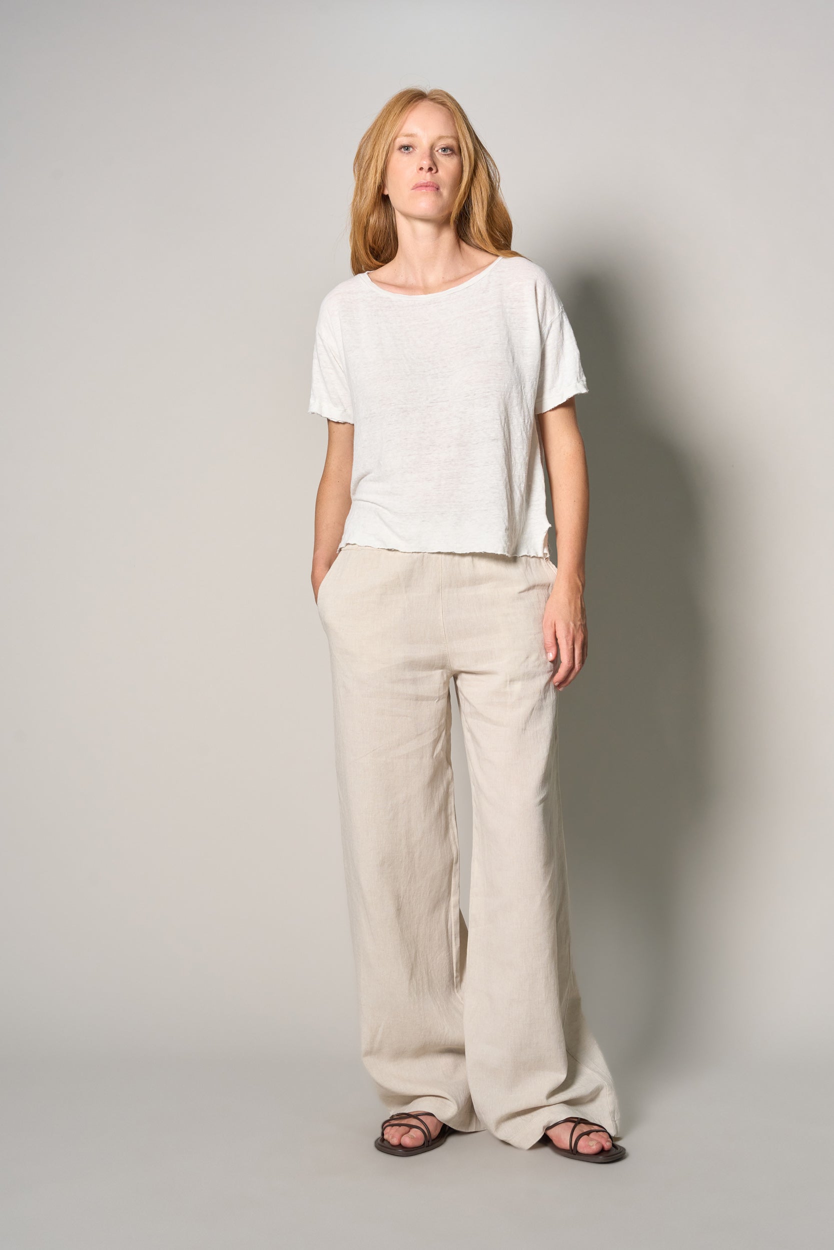 A woman modeling a white short-sleeve crewneck t-shirt and beige wide-leg linen pants, paired with dark sandals, against a neutral light gray studio background.