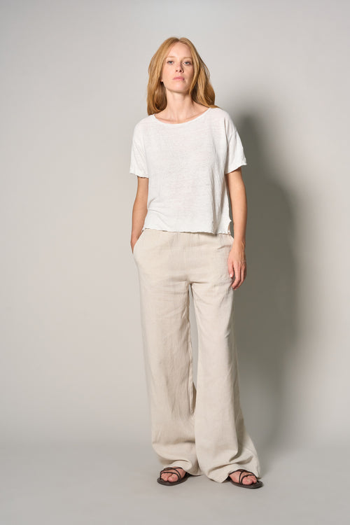 A woman modeling a white short-sleeve crewneck t-shirt and beige wide-leg linen pants, paired with dark sandals, against a neutral light gray studio background.