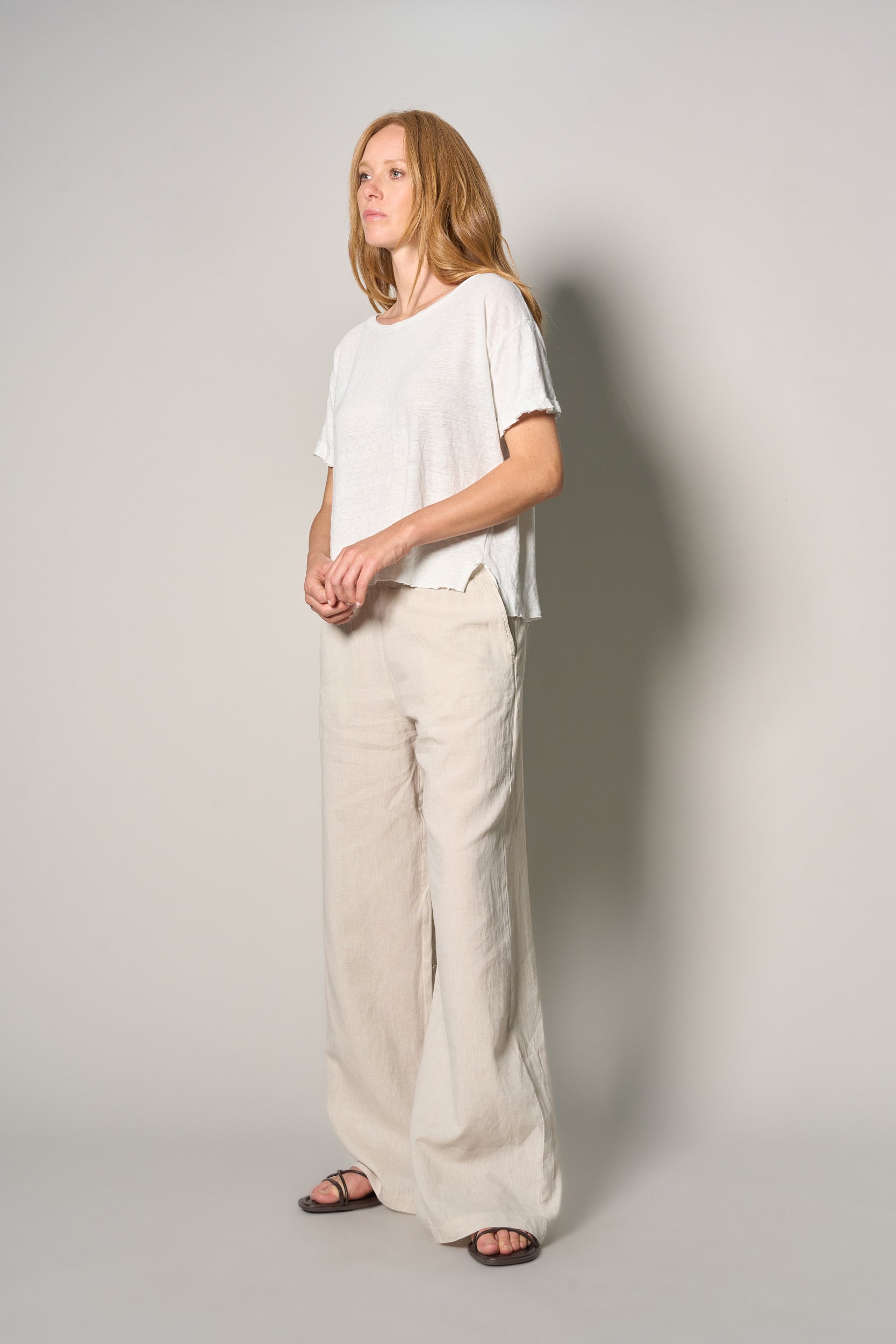 A woman modeling a white short-sleeve top and wide-leg beige trousers against a plain light gray studio background.