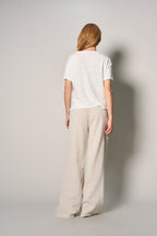 A woman viewed from behind wearing a white short-sleeve t-shirt and light beige wide-leg trousers, standing against a neutral light gray studio background.