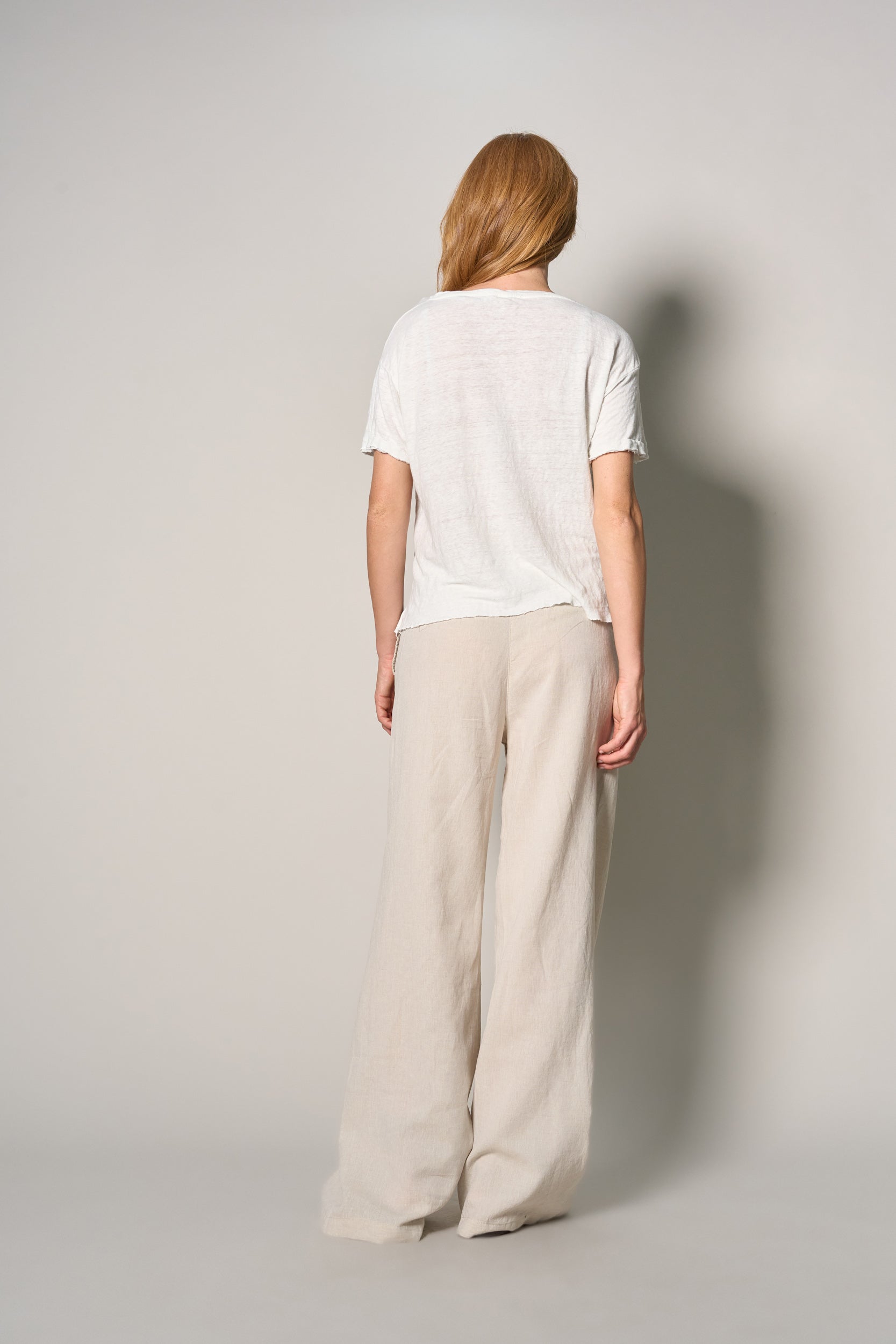 A woman viewed from behind wearing a white short-sleeve t-shirt and light beige wide-leg trousers, standing against a neutral light gray studio background.