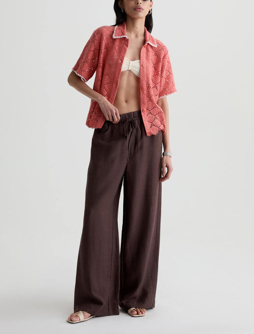 A model wearing a coral pink short-sleeve button-up shirt with a snakeskin pattern and dark brown wide-leg pants, styled with a white bikini top and flat sandals against a light gray studio background.