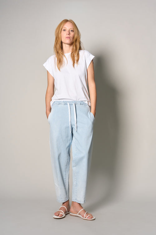 A woman modeling a white short-sleeve t-shirt and light-wash blue relaxed-fit jeans with a white drawstring, paired with white strappy flat sandals, against a neutral gray studio background.