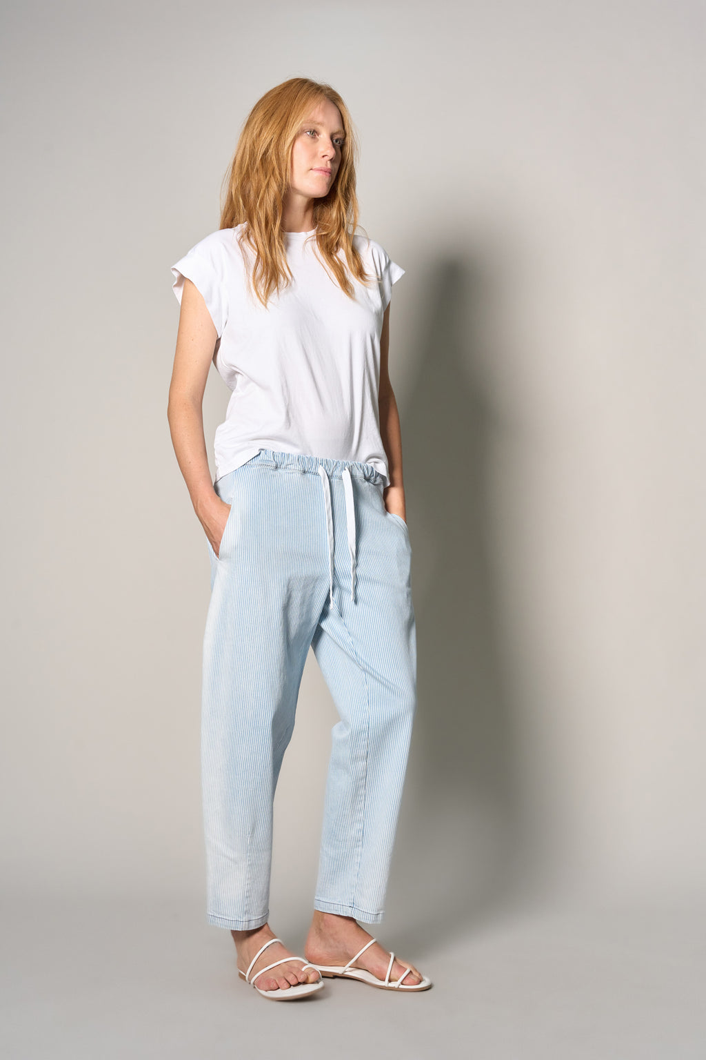 A woman modeling a white short-sleeve t-shirt and light-wash blue drawstring pants, paired with white strappy sandals, standing against a neutral gray studio background.