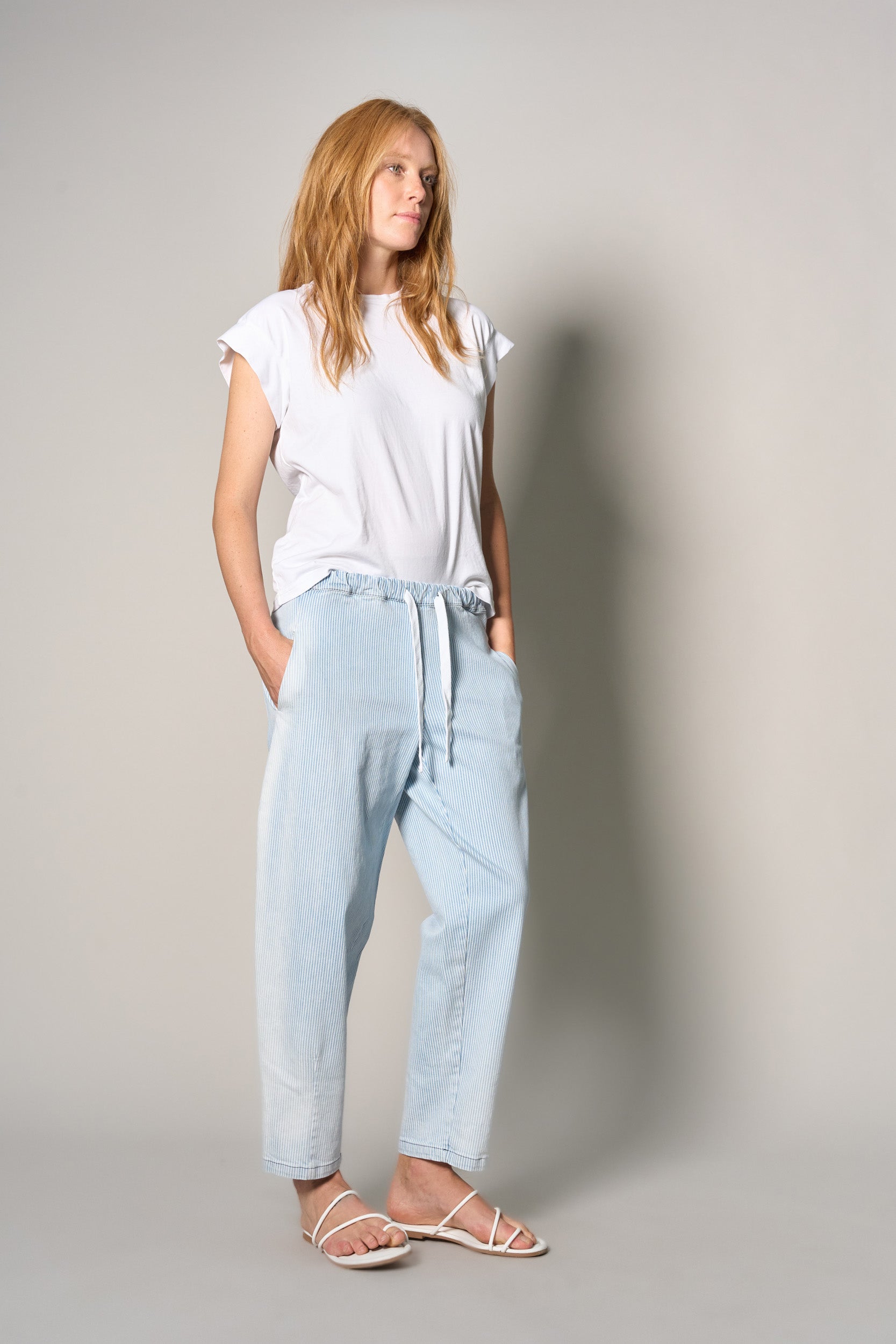 A woman modeling a white short-sleeve t-shirt and light-wash blue drawstring pants, paired with white strappy sandals, standing against a neutral gray studio background.
