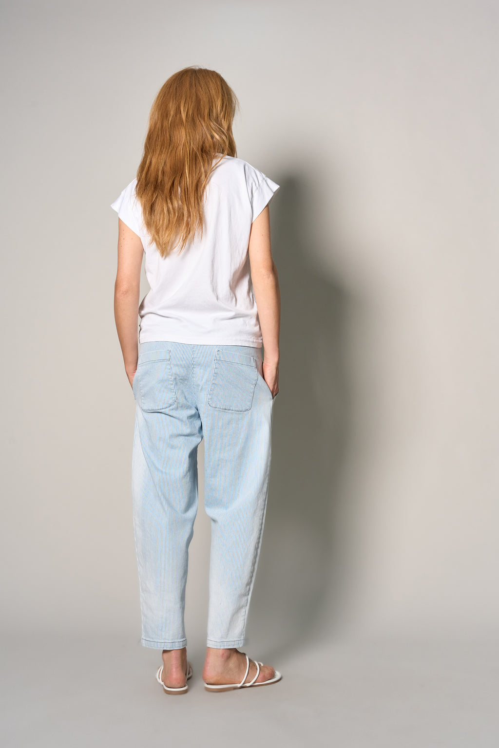 A woman with long red hair is shown from behind wearing a white t-shirt, light blue relaxed-fit jeans, and white flat sandals against a plain light gray studio background.