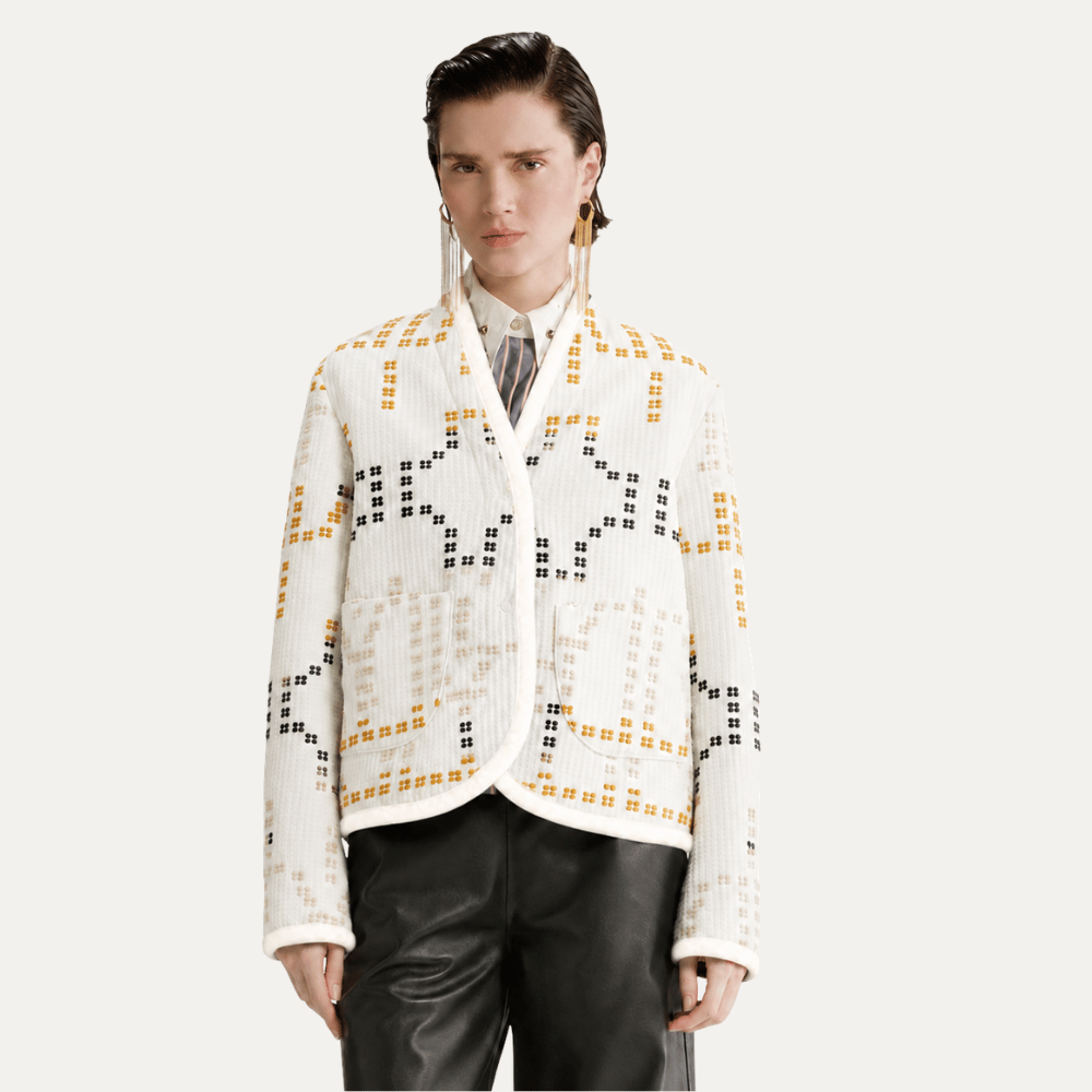 A model wearing a white textured knit cardigan with a geometric black and mustard yellow pattern, styled with black leather pants against a light gray studio background.