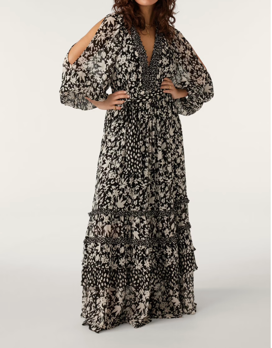 A model wearing a long floral maxi dress with puff sleeves, a V-neckline, and ruffles. The dress has a tie belt and is predominantly black with a pattern of smaller black elements.