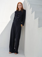 A woman standing with one hand on a staircase, wearing mid waist navy pajama trousers with a mid-rise waist and a slightly flared straight leg, paired with a black blazer with shoulder pads and a gold watch.