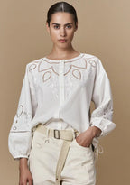 A model wearing a white long-sleeved blouse with floral embroidery and mother-of-pearl buttons, paired with light-colored trousers.