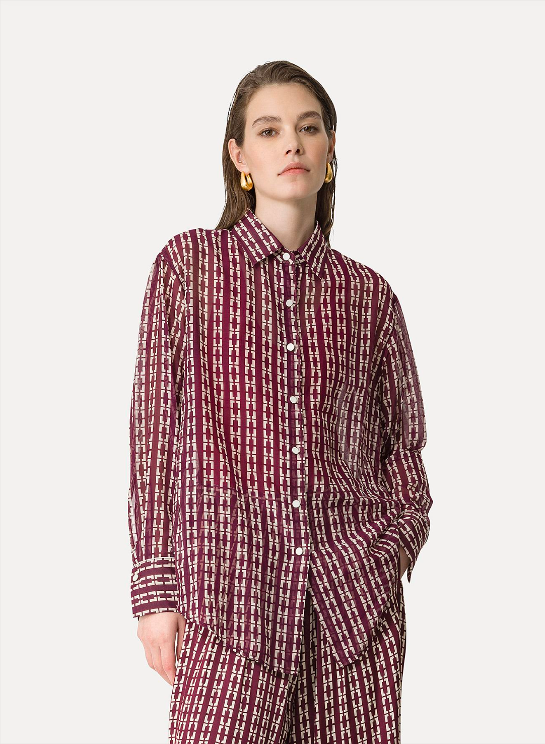A woman wearing a maroon and white geometric patterned oversized shirt with a collar and buttons down the front, paired with matching pants.