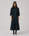 A woman standing wearing a navy blue dress with a belted waist, cap off, and a hat on. The dress has a collared neckline and is designed for a flattering fit.