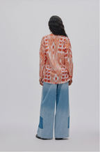 A model viewed from behind wearing a long-sleeve button-up shirt with an orange and white abstract pattern, paired with wide-leg light blue jeans, against a solid light gray background.