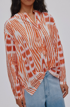 A woman wearing a long-sleeve button-front shirt with an orange and white tie-dye pattern, paired with light blue jeans, shown against a plain light gray background.