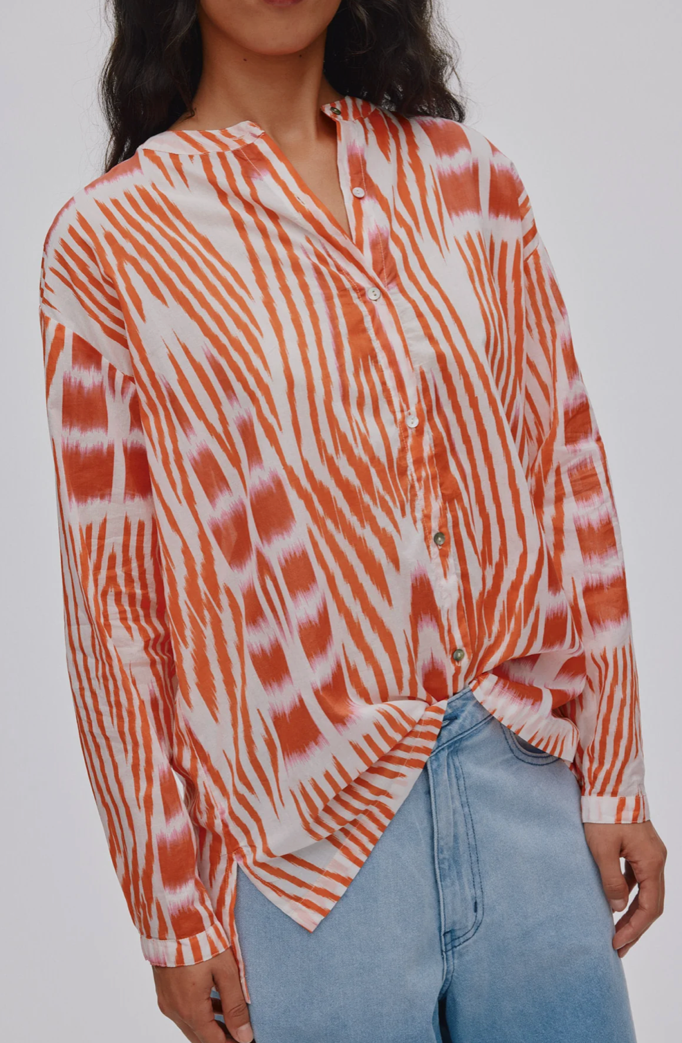 A woman wearing a long-sleeve button-front shirt with an orange and white tie-dye pattern, paired with light blue jeans, shown against a plain light gray background.