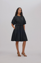 A woman modeling a black short-sleeve midi dress with a gathered waist and pleated skirt, paired with brown strappy high-heeled sandals, against a light gray studio background.