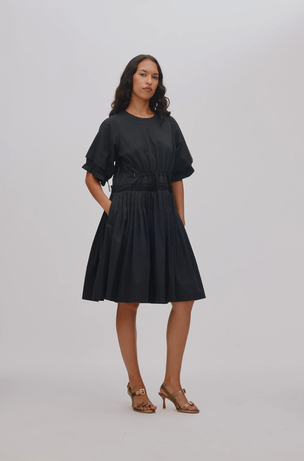 A woman modeling a black short-sleeve midi dress with a gathered waist and pleated skirt, paired with brown strappy high-heeled sandals, against a light gray studio background.