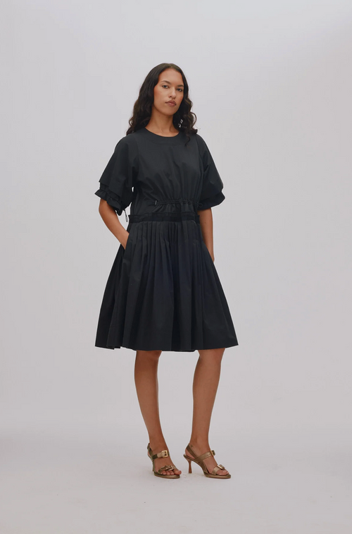 A woman modeling a black short-sleeve midi dress with a gathered waist and pleated skirt, paired with brown strappy high-heeled sandals, against a light gray studio background.