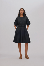 A woman modeling a black midi dress with short puff sleeves, a gathered waist, and a pleated skirt, paired with brown heeled sandals against a light gray studio background.