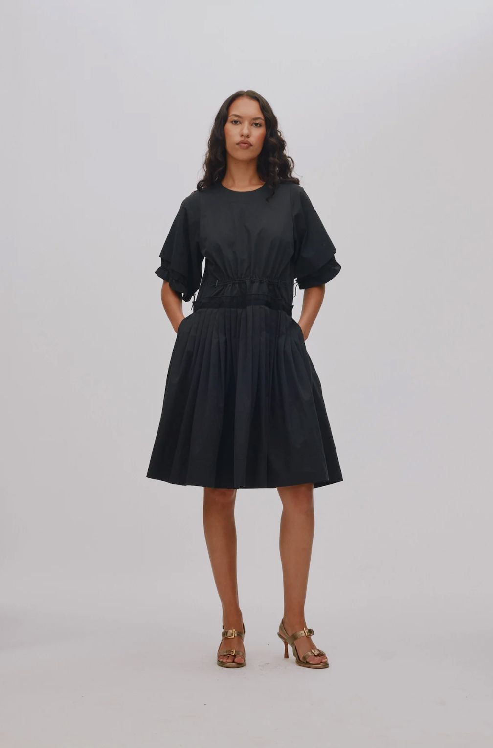 A woman modeling a black midi dress with short puff sleeves, a gathered waist, and a pleated skirt, paired with brown heeled sandals against a light gray studio background.