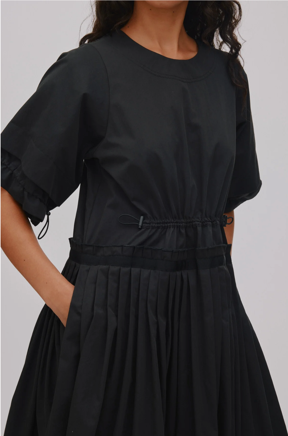 A close-up of a model wearing a black short-sleeve dress with a gathered waist, pleated skirt, and side tie details, set against a plain light gray background.