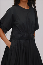 A close-up of a model wearing a black short-sleeve dress with a gathered waist, pleated skirt, and side tie details, set against a plain light gray background.
