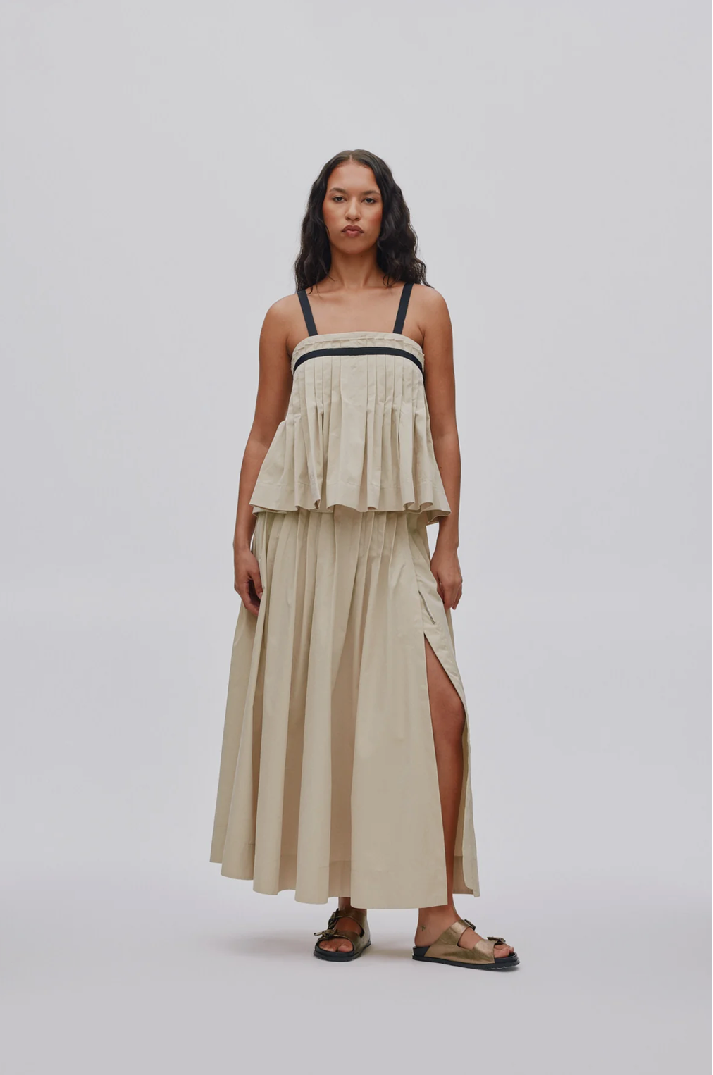 A woman modeling a light beige sleeveless maxi dress with a tiered peplum top, thin black shoulder straps, and a side slit, paired with brown sandals, against a light gray studio background.