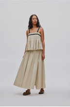 A woman modeling a long cream-colored sleeveless maxi dress with a layered ruffle bodice and thin black shoulder straps, paired with brown leather sandals, against a light gray studio background.